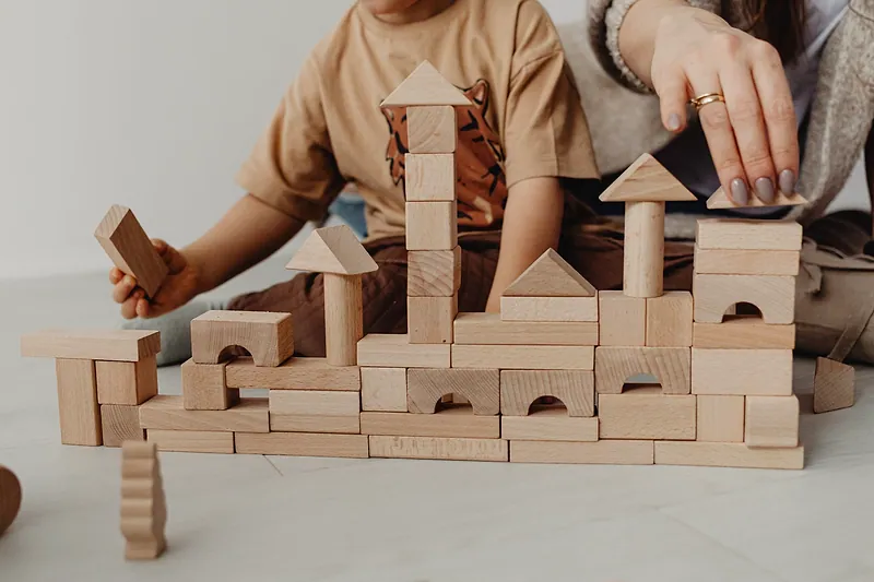 La mano de un niño y la de una persona adulta construyen un castillo de bloques de madera.
